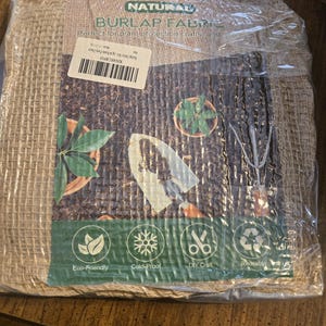 May include: A square package of natural burlap fabric wrapped in clear plastic. The label features the text NATURAL BURLAP FABRIC, images of a garden trowel and plants, and four circular icons labeled Eco-Friendly, Cold-Proof, DIY Craft, and Reusable.
