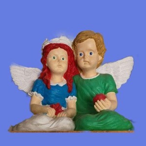 May include: A figurine of two angels, one with red hair and a white crown, the other with brown hair. The angel with red hair is wearing a blue dress and white wings. The angel with brown hair is wearing a green shirt and white wings. Both angels are holding a red apple.