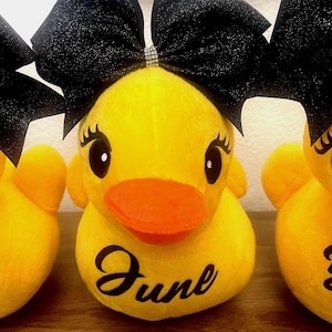 May include: Yellow plush duck with an orange beak and black eyes. The duck has a black glitter bow with a rhinestone center. The word "June" is written in black script on the duck's body. The image shows three ducks.