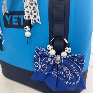 May include: A bright blue cooler bag with a black top and bottom trim, featuring a YETI logo. Attached to the side is a blue bandana-print fabric bow with a rhinestone centre, suspended by a beaded loop with black and white football beads.