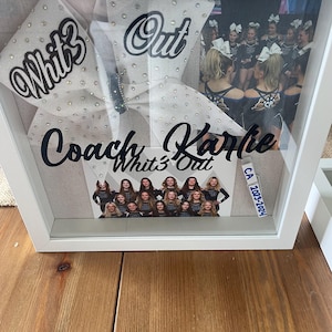 Puede incluir: Una caja de sombra enmarcada en blanco con un lazo de animadora con las palabras "Whit3 Out" y "Out" en escritura negra. La caja también contiene fotos del equipo y el nombre "Coach Karlie". El año "2023-2024" también es visible.