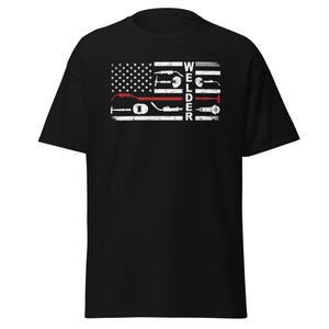 May include: Black t-shirt featuring a distressed graphic of the American flag with welding tools and the word "WELDER" in white. The design includes a red stripe, symbolizing the thin red line. A patriotic and occupational themed apparel.