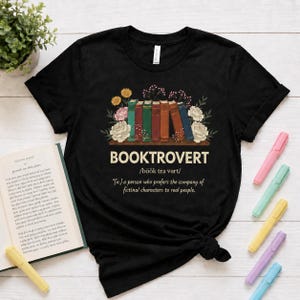 May include: A black short-sleeved t-shirt featuring a graphic of five colorful books arranged in a row with floral accents. Below the books, the text reads BOOKTROVERT /bʊk trə vɜrt/ *[n.] a person who prefers the company of fictional characters to real people.