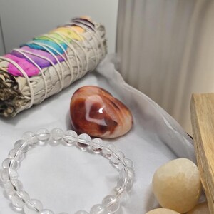 Peut inclure: Une collection d'articles spirituels comprenant un bracelet en quartz clair, un fagot de sauge avec des plumes aux couleurs de l'arc-en-ciel, une pierre d'agate polie et plusieurs pierres beiges lisses. Un bâton de bois est également présent.