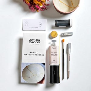 Pode incluir: Um kit de reparação Kintsugi com uma tigela de cerâmica quebrada, resina epóxi dourada, pincéis e ferramentas. O kit inclui um manual intitulado "Kintsugi Moderno" e um buquê de flores secas. A tigela tem detalhes dourados.