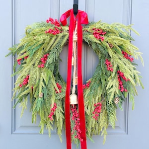 May include: A festive Christmas wreath with green faux pine needles, red berries, and small pinecones. A red satin ribbon and a jute rope with a small bell hang from the top. The wreath is displayed on a gray door.