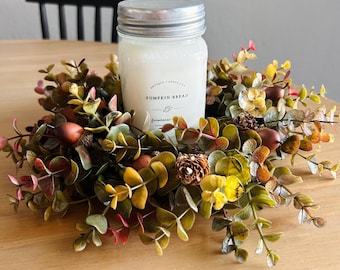 May include: A jar candle with a silver lid, labeled "Pumpkin Bread," sits in a decorative wreath. The wreath features green, yellow, and red leaves, small pinecones, and acorns. The candle and wreath are on a wooden table.