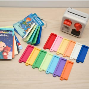 以下が含まれることがあります： オレンジ色のノブが付いた白いオーディオプレーヤー、ココメロンの子守唄カードの束、そして赤、ピンク、黄色、オレンジ、白、青、緑、ライトグリーン、ライトブルー、紫、ダークオレンジのカラフルな長方形のプラスチックフレームのコレクション。