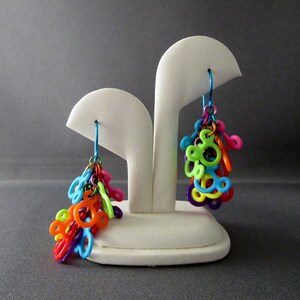 Puede incluir: Un par de pendientes colgantes de colores con forma de Mickey Mouse. Los pendientes est&aacute;n hechos de pl&aacute;stico y cuelgan de un expositor blanco.