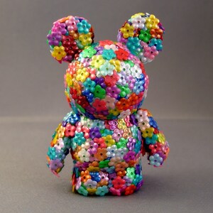 Op de afbeelding: Een kleurrijk speelgoedfiguur bedekt met kleine, veelkleurige bloemen. De figuur heeft een rond hoofd met grote oren en een klein lichaam. De bloemen zijn in een willekeurig patroon gerangschikt en cre&euml;ren een levendig en vrolijk ontwerp.