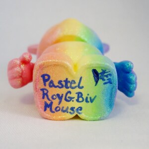 May include: A rainbow-coloured mouse figurine with glitter. The mouse is standing on its hind legs with its front paws outstretched. The mouse is signed "Pastel RoyG. Biv Mouse".
