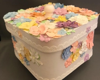 Può includere: Una scatola quadrata bianca con un coperchio dotato di pomello centrale e una fitta disposizione di fiori di carta color pastello nelle tonalità rosa, giallo, blu e viola. I lati sono decorati con bordi in pizzo bianco e fiori aggiuntivi.