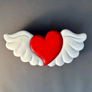 Puede incluir: Un corazón rojo con alas blancas. El corazón está pintado en un estilo 3D y las alas están pintadas en un estilo plano. El corazón está centrado en la imagen y las alas están extendidas a ambos lados del corazón.