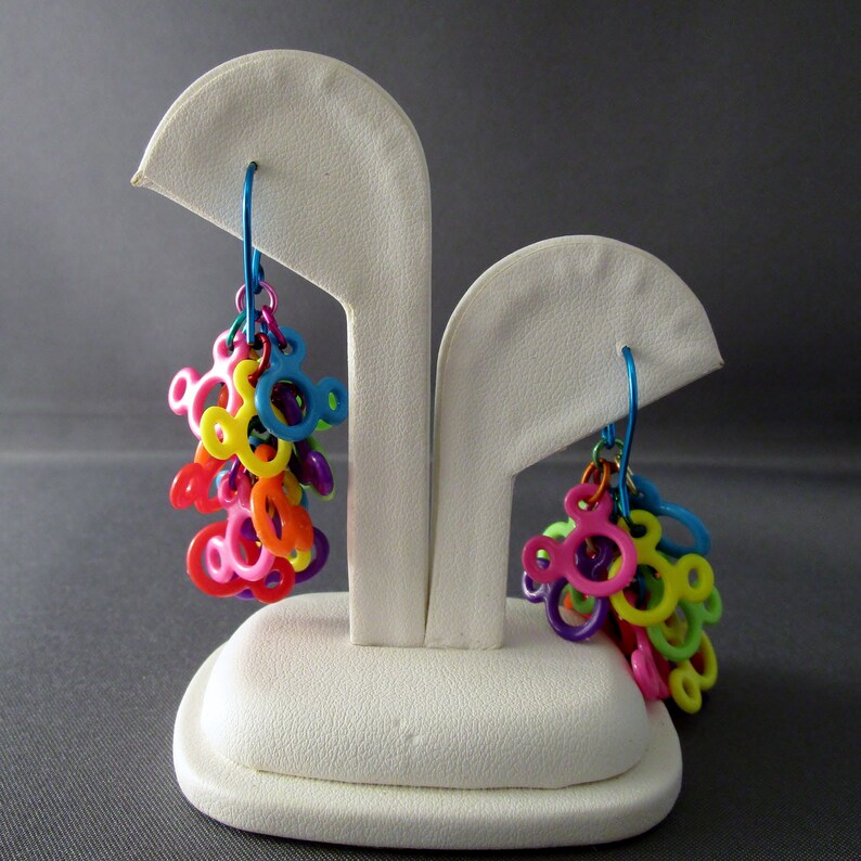 Puede incluir: Un par de pendientes colgantes de colores con un dise&ntilde;o de cabeza de Mickey Mouse. Los pendientes est&aacute;n hechos de pl&aacute;stico y est&aacute;n enhebrados en un alambre plateado. Los pendientes se muestran en un soporte de pendientes blanco.