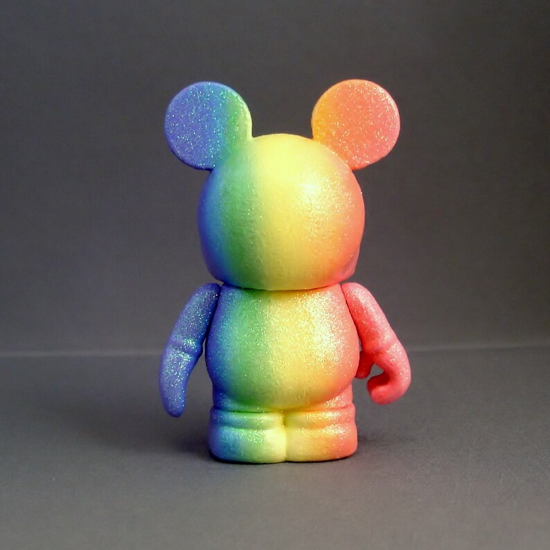 May include: A rainbow-coloured, glittery, vinyl toy figure with a Mickey Mouse design. The figure has a round head with large ears and a small body with short legs and arms.