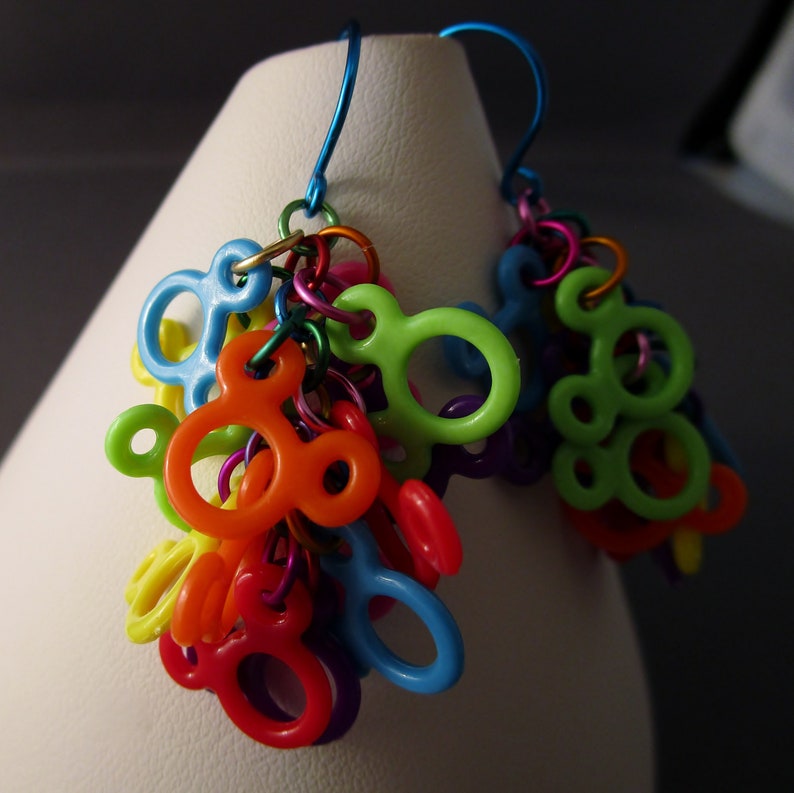 Puede incluir: Un par de pendientes colgantes coloridos con m&uacute;ltiples dijes en forma de Mickey Mouse en varios colores, incluyendo verde, naranja, azul, rojo y amarillo. Los dijes est&aacute;n conectados por eslabones de cadena plateados.