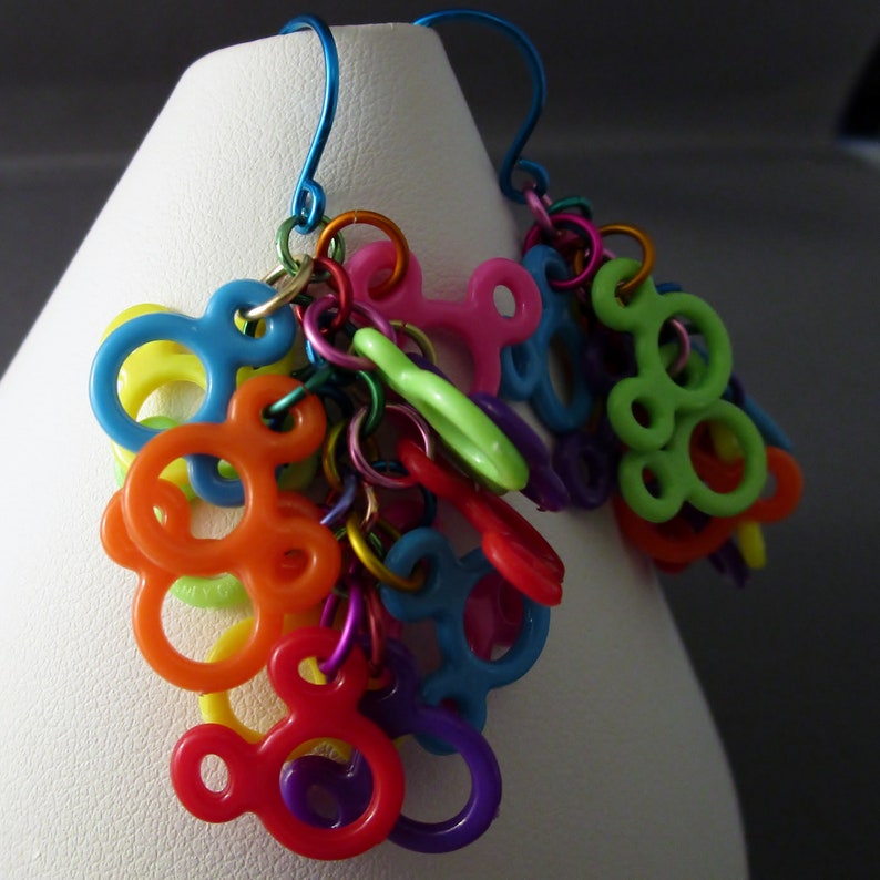 Puede incluir: Pendientes colgantes coloridos con m&uacute;ltiples dijes de pl&aacute;stico con forma de orejas de Mickey Mouse. Los dijes est&aacute;n unidos por una cadena plateada y cuelgan de ganchos plateados.
