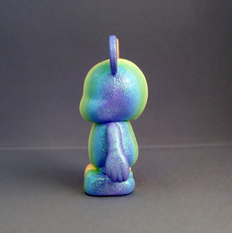 May include: A small, blue, green and purple figurine with a glittery finish. The figurine is shaped like a bear with large ears and a rounded body.