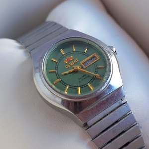 May include: A silver-tone wristwatch with a round green dial, gold-coloured hour markers, and gold hands. The watch features a day-date window at the three o'clock position, the Orient logo, and a metallic link bracelet, presented in a box.