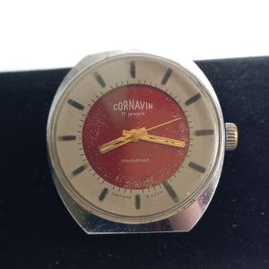 May include: A silver-toned wristwatch with a round face featuring a deep red center and a light-colored outer ring. The dial displays the text CORNAVIN, 17 jewels, and shockproof, with gold-toned hands and black hour markers. A crown is on the side.