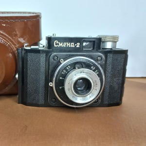 Peut inclure: Un appareil photo argentique noir Smena-2 avec un objectif argenté doté de marquages d'ouverture et un étui de transport en cuir marron avec un logo circulaire en relief sur le devant. Le boîtier a une finition texturée et une molette argentée.