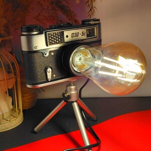 Può includere: Una lampada da scrivania unica realizzata con una fotocamera vintage, con una grande lampadina in stile Edison. La fotocamera, nera e argento, è montata su un piccolo treppiede. La lampadina emette una luce calda, ideale per un ufficio domestico o uno spazio creativo.