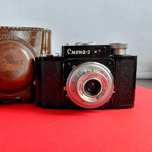 Peut inclure: Un appareil photo argentique noir Smena-2 avec un objectif argenté et un étui de transport en cuir marron. Le boîtier de l'appareil porte l'inscription cyrillique "Смена-2" sur le devant. L'objectif comporte des bagues métalliques avec des graduations.