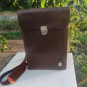 May include: A brown leather satchel with a flap closure and a shoulder strap. The bag has a silver-colored clasp and a small decorative emblem on the front. The satchel is sitting on a white surface.