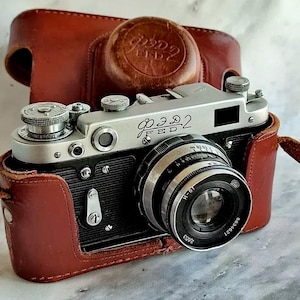 May include: A vintage silver and black FED 2 camera with a lens, resting in a brown leather case. The camera has several dials and a lens with a black and silver design. The leather case has a matching lens cap.