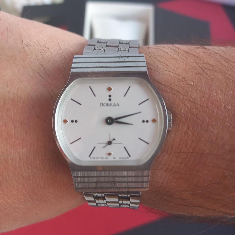 May include: A vintage silver-toned wristwatch with a white face and black hands. The watch face features the word "ПОБЕДА" and small markers. The watch is on a silver-toned metal band.