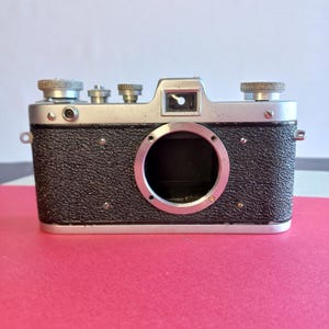 以下が含まれることがあります： 黒とシルバーのヴィンテージカメラ。テクスチャ加工されたボディと複数のダイヤルがあります。カメラには大きな円形のレンズ開口部とビューファインダーがあります。背景はピンクと白です。