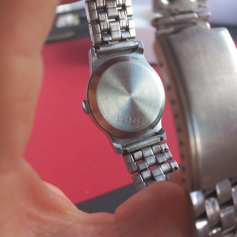 May include: Close-up of a silver-toned wristwatch. The watch has a round, brushed metal back with the number "194345" etched into it. The watch band is a linked design.