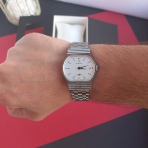 May include: A silver-toned wristwatch with a white face and a rectangular case. The watch has a silver band and is worn on a wrist. The watch face has black hands and small red accents. The brand name "DONGIA" is printed on the watch face.