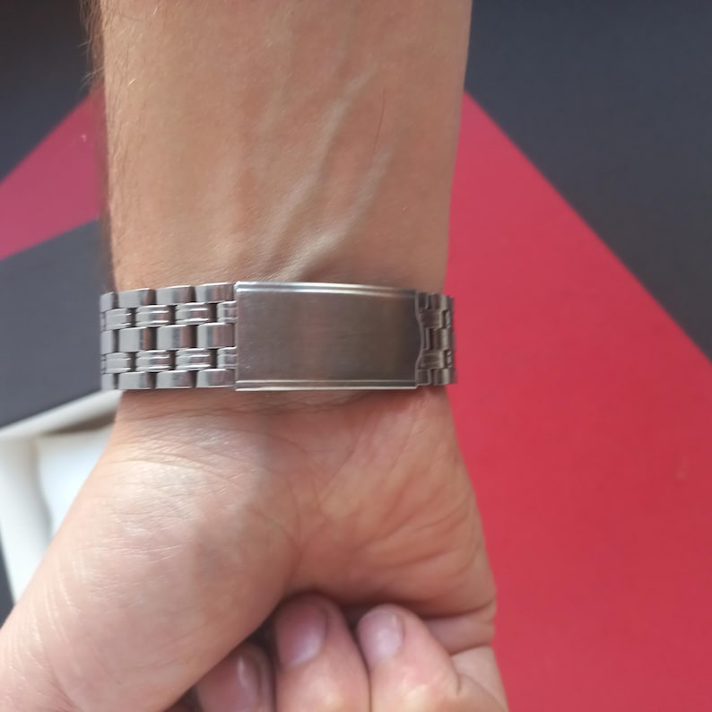 May include: A silver-toned watch band with a rectangular clasp is fastened on a wrist. The band is made of small, rectangular links. The clasp is a solid, brushed metal. The background is a mix of red, black, and white.
