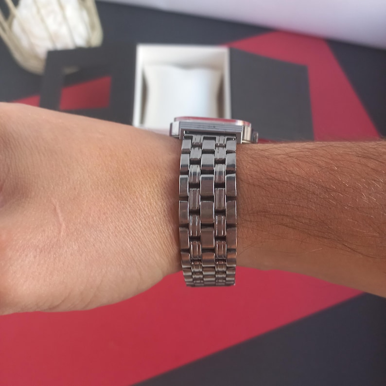 May include: A silver wristwatch with a rectangular face and a linked metal band. The watch is on a wrist, with the band wrapping around the arm. The watch face is red. The watch is in front of a black and red surface.