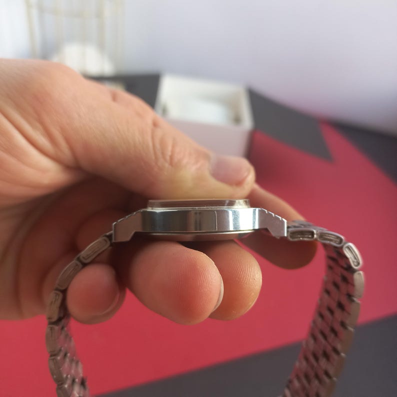 May include: A silver-toned wristwatch with a rectangular face and a linked metal band. The watch is held in a hand, showcasing the side profile of the watch and band. The watch face appears to be a light color, and the band is made of small, connected metal pieces.