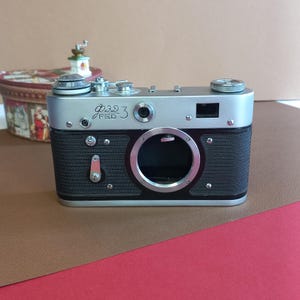 Puede incluir: Una cámara FED 3 vintage con la parte superior plateada y el cuerpo negro. La cámara tiene una abertura prominente para la lente y varios diales y botones. El texto "FED 3" es visible en la parte delantera. Sobre un fondo marrón y rojo.