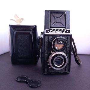 Peut inclure: Un appareil photo reflex bi-objectif Lubitel 2 noir avec une sangle en cuir, un bouchon d'objectif et un étui. L'appareil photo possède deux objectifs circulaires et le nom "Lubitel 2" est imprimé sur le devant. Une petite figurine d'oiseau blanc est posée sur l'étui.