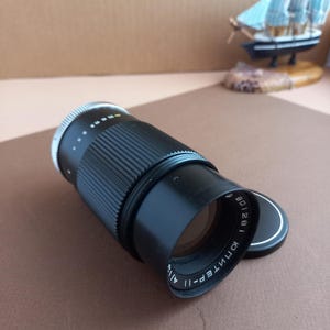 Op de afbeelding: Een zwarte cameralens met een zilveren instelring en een zwarte zonnekap. De lens heeft witte markeringen en een zwarte lensdop met witte tekst. De lens ligt op een bruin oppervlak.