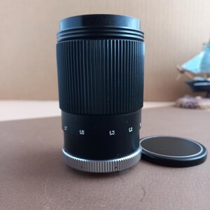 Op de afbeelding: Een zwarte cameralens met een geribbeld lichaam en een zilveren basis. De lens heeft witte markeringen die brandpuntsafstanden aangeven. Een zwarte lensdop ligt ernaast. De lens is ontworpen voor fotografie.