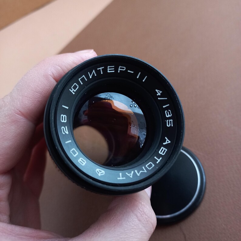 Op de afbeelding: Een zwarte cameralens met de tekst "ЮПИТЕР-11" en "4/135 ABTOMAT" in witte letters. De lens heeft een reflecterend glazen oppervlak en een zwarte lensdop.