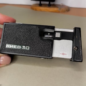 May include: A black Kiev-30 subminiature camera with a textured body and a sliding silver metal panel. The front features the text KIEV-30 in white lettering, a small silver shutter button, a focus dial, and a red circular symbol.