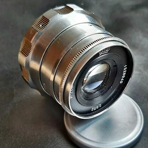 May include: A silver-toned metal camera lens with a knurled focus ring and a black front element. The lens features the text 2.8/52 and the serial number 1528940 printed on the front. A matching silver metal lens cap is positioned underneath the lens.