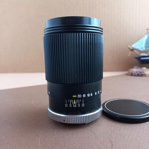 Op de afbeelding: Een zwarte cameralens met geribbelde zijkanten en een zilveren basis. De lens heeft markeringen, waaronder diafragma's en afstands schalen. Een zwarte lensdop ligt ernaast.