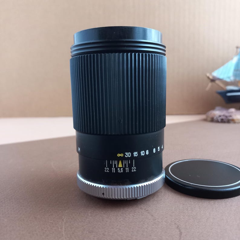 Op de afbeelding: Een zwarte cameralens met geribbelde zijkanten en een zilveren basis. De lens heeft markeringen, waaronder diafragma's en afstands schalen. Een zwarte lensdop ligt ernaast.