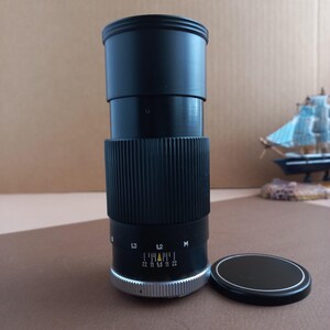 Op de afbeelding: Een zwarte cameralens met een geribbelde grip en een zilveren ring aan de basis. De lens heeft f-stop markeringen en een zwarte lensdop. Een miniatuur zeilboot staat op de achtergrond.