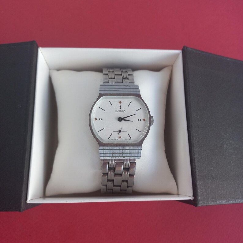 May include: A vintage silver-toned wristwatch with a white face, black hands, and the word "ПОБЕДА" printed on the dial. The watch is in a black and white box, resting on a white pillow. The watch band is also silver-toned.