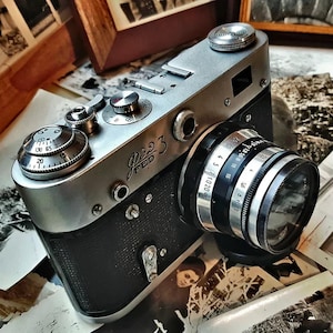 May include: A silver and black FED 3 rangefinder camera featuring a textured black body, a metallic top plate with circular dials, and a lens with engraved aperture and focus markings. The camera body displays the text FED 3 in a stylized font.