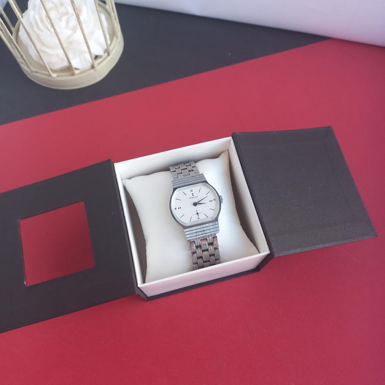 May include: A silver-toned wristwatch with a white face and a rectangular case is displayed in an open black box with a white interior. The watch has a linked metal band. The box is on a red surface.