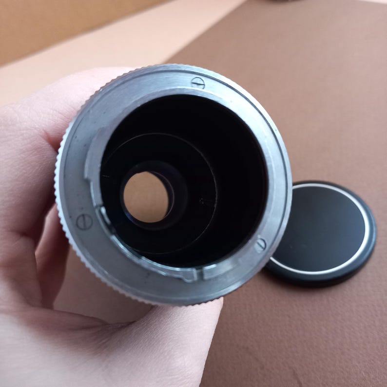 Op de afbeelding: Een zilverkleurige camera lens adapter met een zwart interieur en een zwarte lensdop. De adapter heeft een geribbelde rand voor grip en diverse kleine schroeven. De lensdop is rond met een zilveren rand.